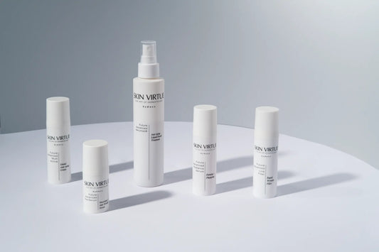 Six white skincare containers from the brand SKIN VIRTUE are arranged on a light gray surface against a matching light gray background. The set includes various sizes of bottles and tubes with minimalist black text.