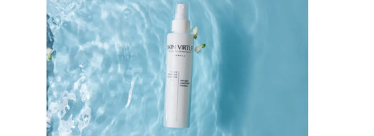 A white spray bottle labelled SKIN VIRTUE Future Advanced Serumist® stands upright against a bright blue water-texture background with bubbles and a small white flower accent.