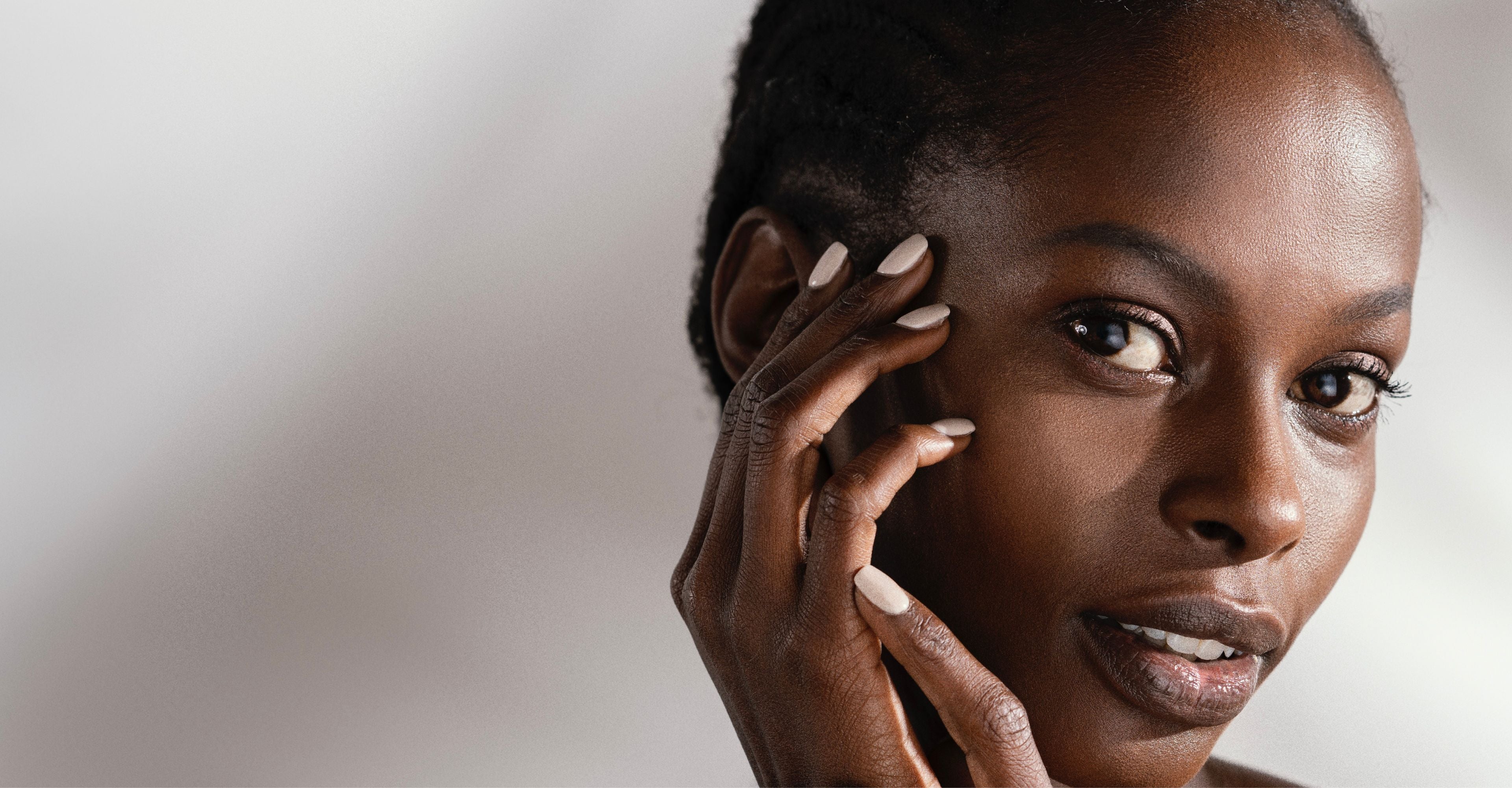 A close-up portrait of a woman with dark skin and short dark hair, touching her face with her hand, set against a soft light gray background.